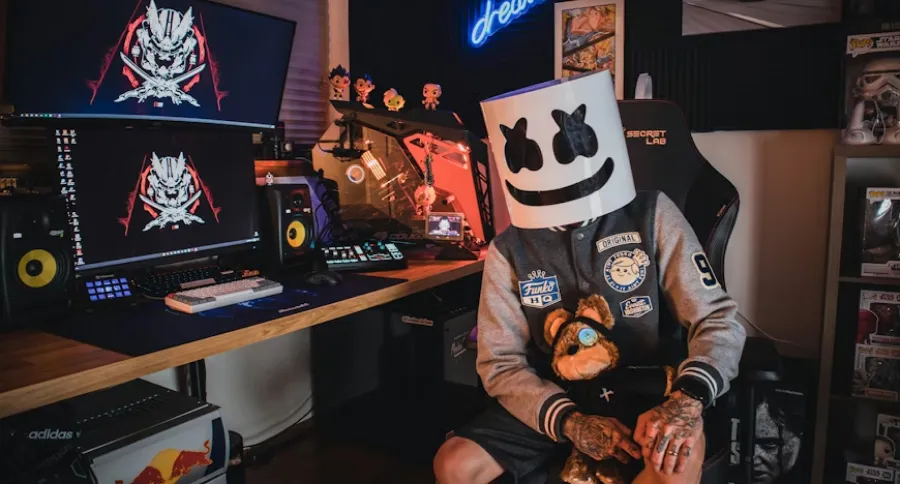 A streamer with a mask sitting in a room equipped with different computers and streaming set up