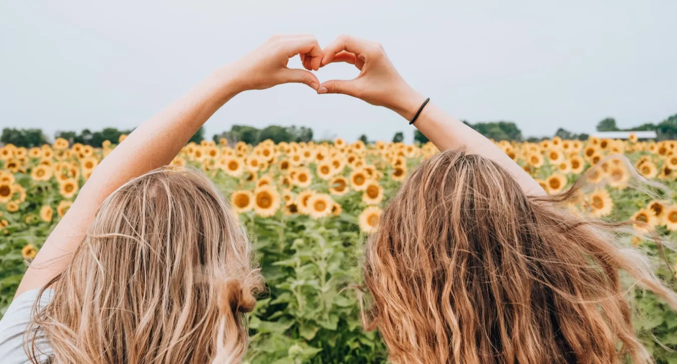 two women in a sunflower field making a heart with their hands together