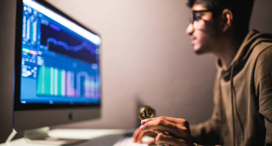 a black haired man with glasses working on a computer while holding a Bitcoin symbol