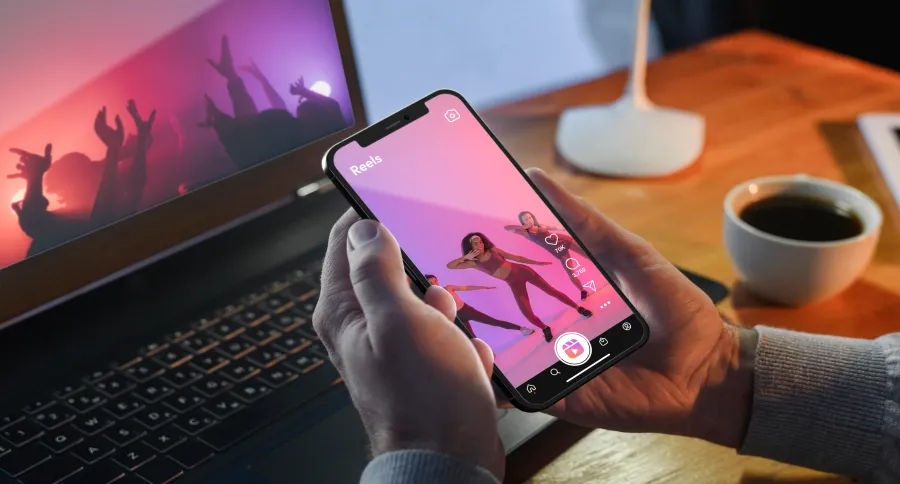 a person watching Instagram Reels on their phone with a laptop on a desk on the background