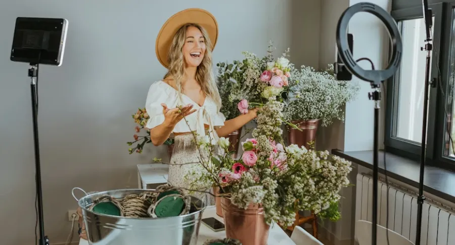 a woman filming in a room filled with flowers with different kind of filming lights and a phone propped up on a holder